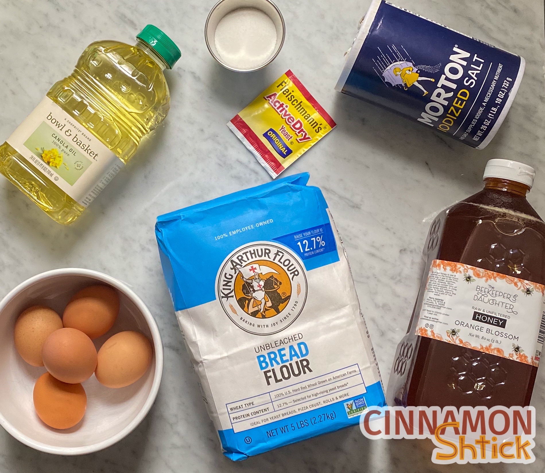 All of the ingredients to make challah: bottle of oil, bowl of eggs, bag of bread flour, bottle of honey, package of salt, package of yeast, and bowl of sugar. 