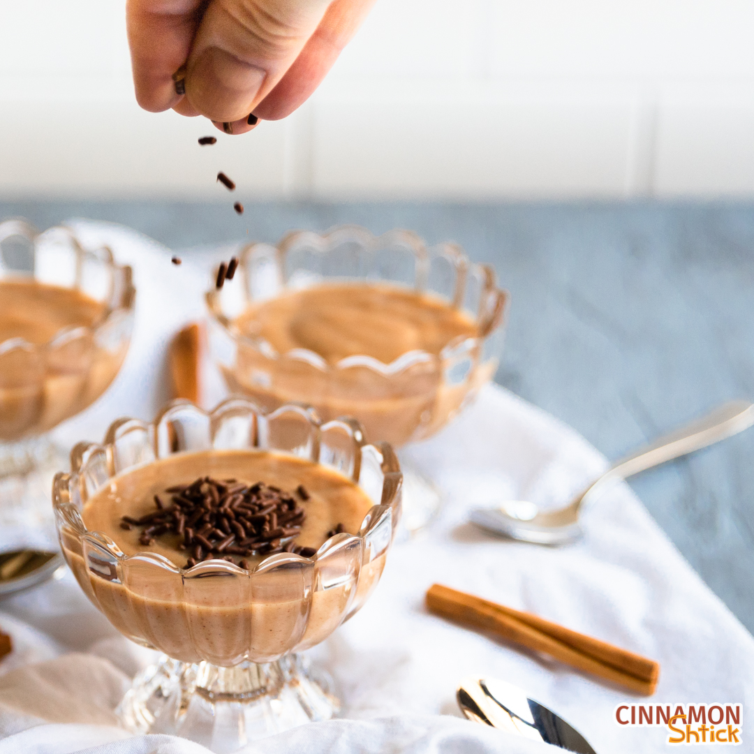 Several dishes of cinnamon pudding on table with spoons and cinnamon sticks with fingers dropping chocolate sprinkles on top of the dish in the forefront.