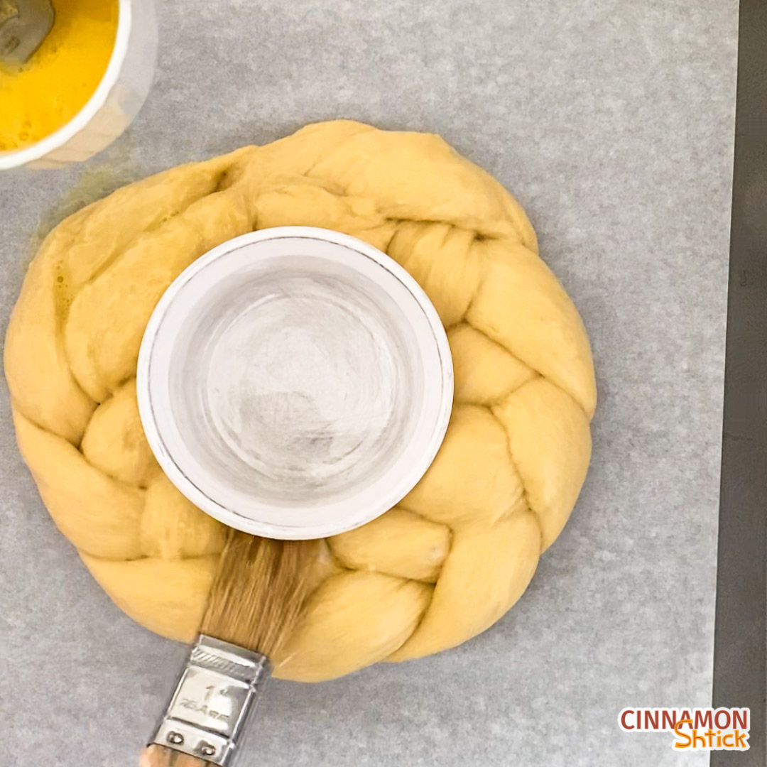 Three strand challah formed in a circle with a ramekin inserted in the middle. Shown with a pastry brush applying egg wash.