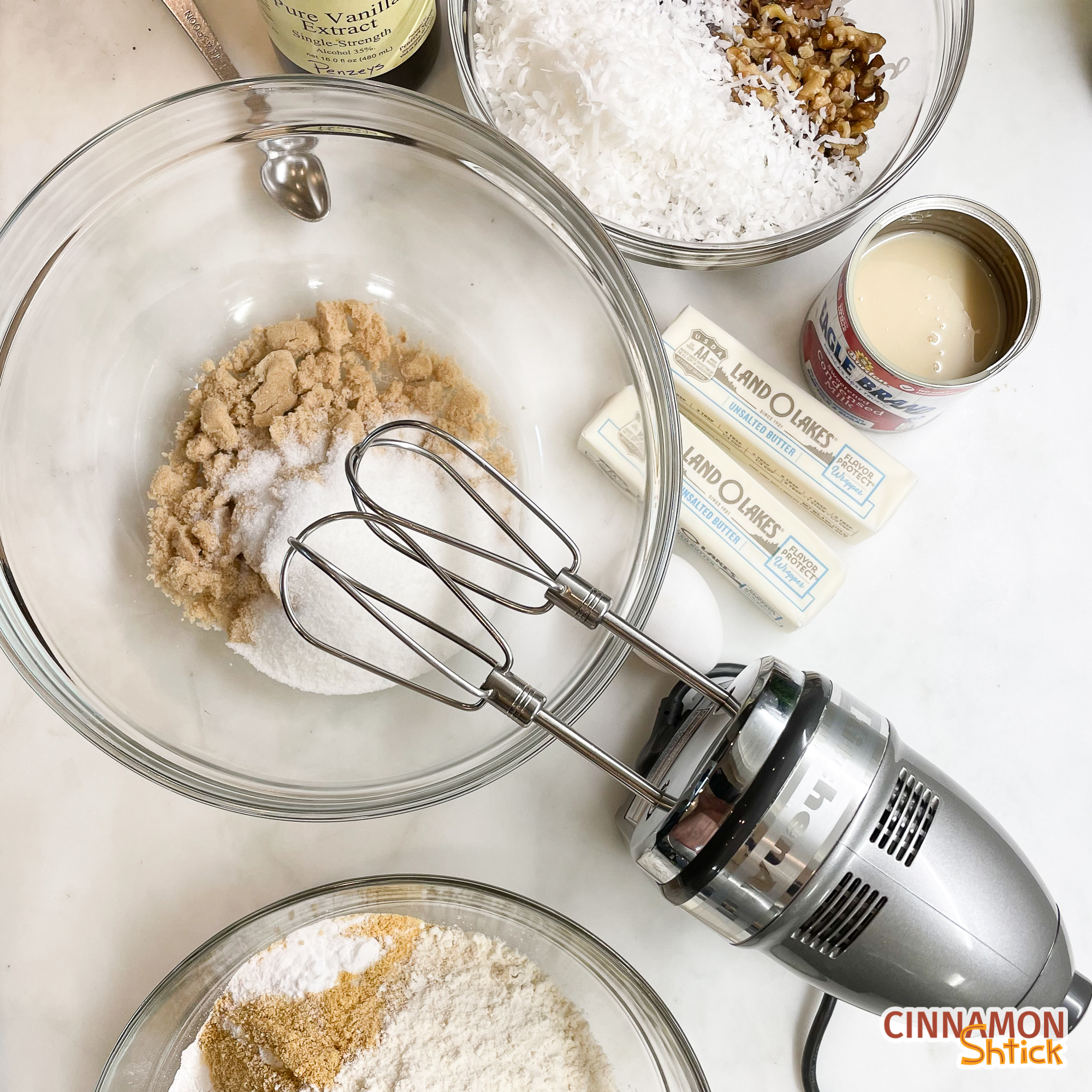 overhead view of hand mixer with bowl of sugars, bowl of dry ingredients, bowl of mix-ins, vanilla, butter, condensed milk