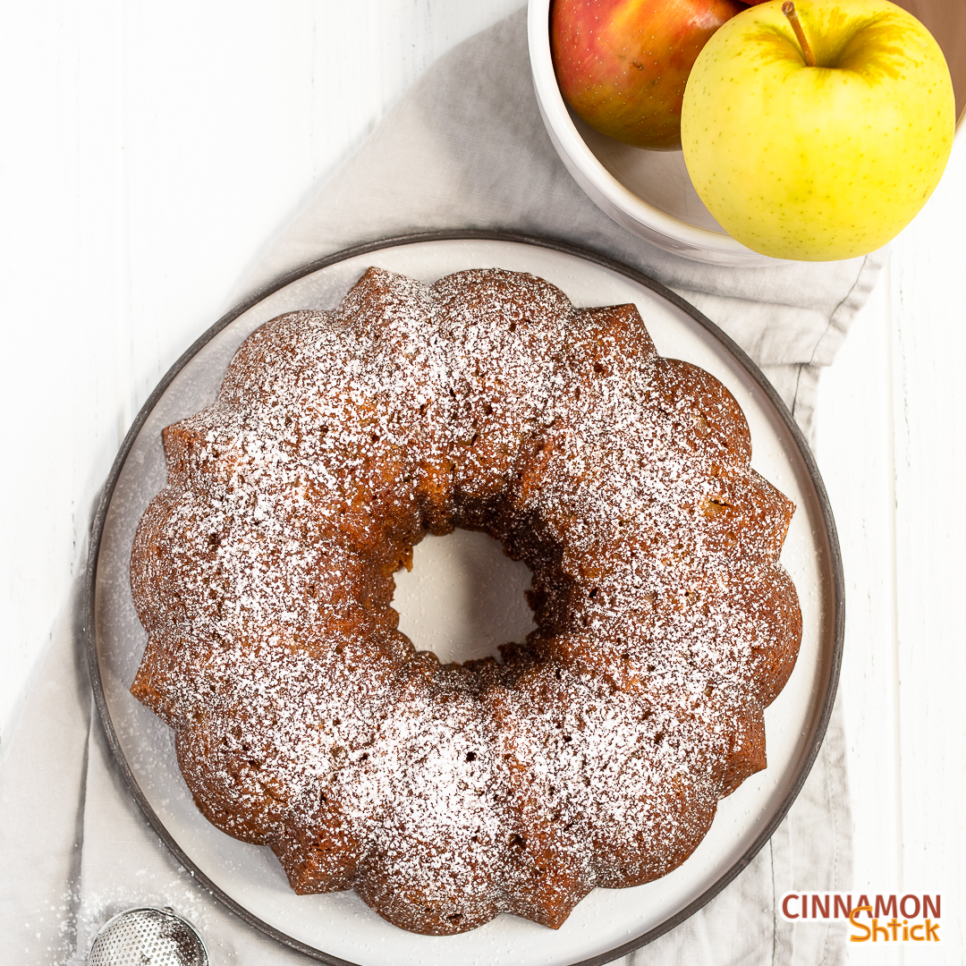 overhead view of apple bundt cake with confectioner's sugar sprinkled on top with a bowl of apples in the upper right corner and a handheld sifter in the bottom left corner