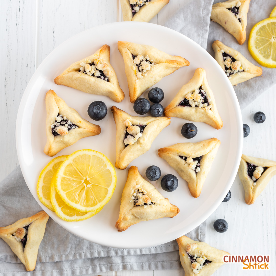 Overhead view of blueberry. hamantaschen on white cake stand with a few slices of lemons and blueberries. Additional blueberry hamantaschen are on tabletop and napkin under the stand.