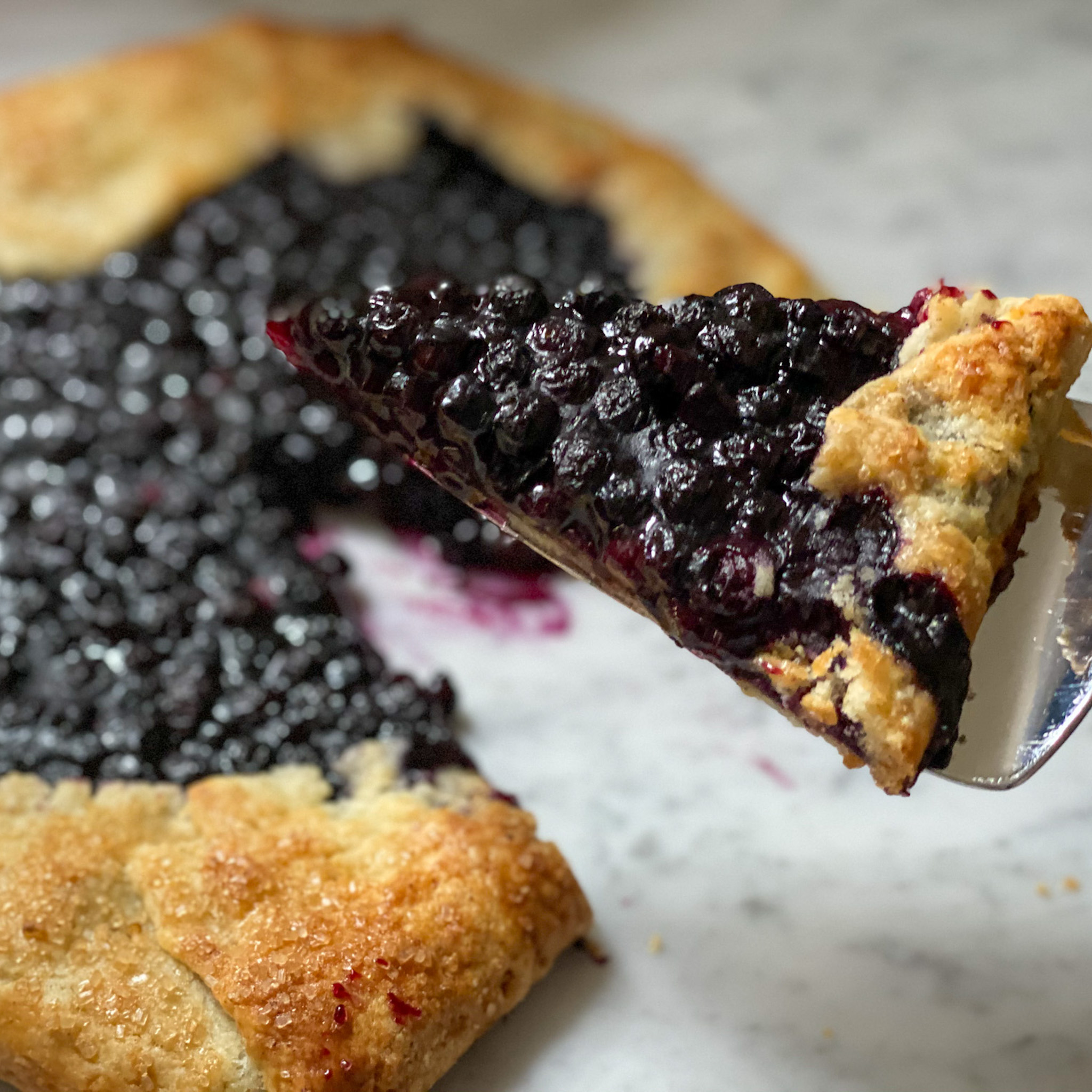 Slice of blueberry galette on a server with rest of galette in background