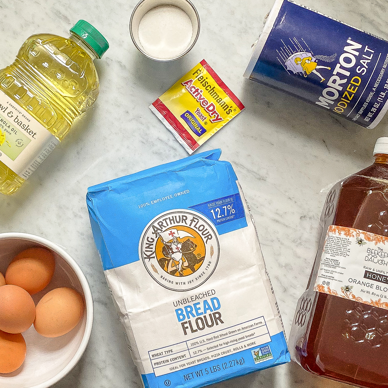 overhead view of ingredients for challah: neutral oil, sugar, yeast, salt, honey, bread flour, and 5 egg yolks.