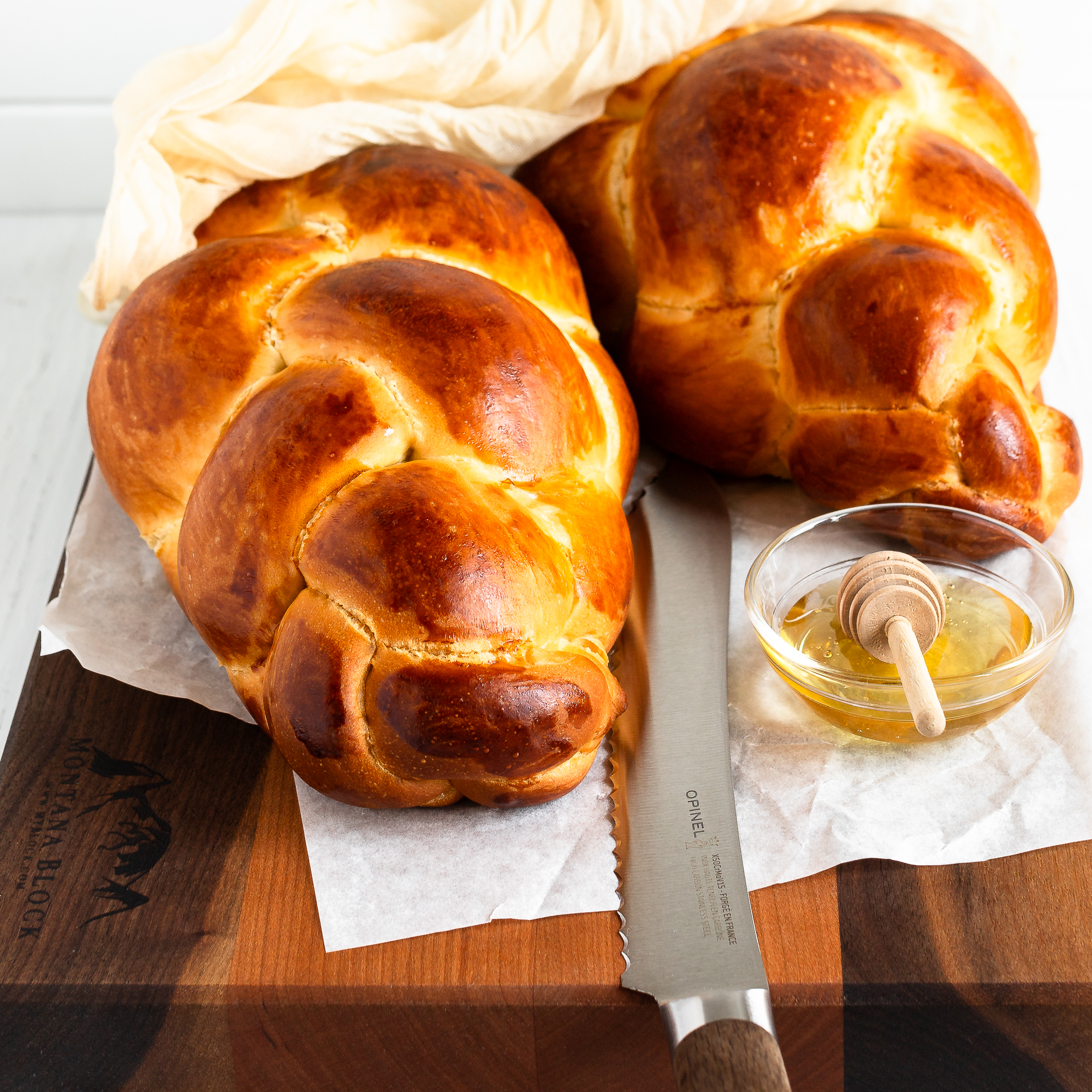 two braided challahs on a cutting board with a bread knife and small bowl of honey in the foreground