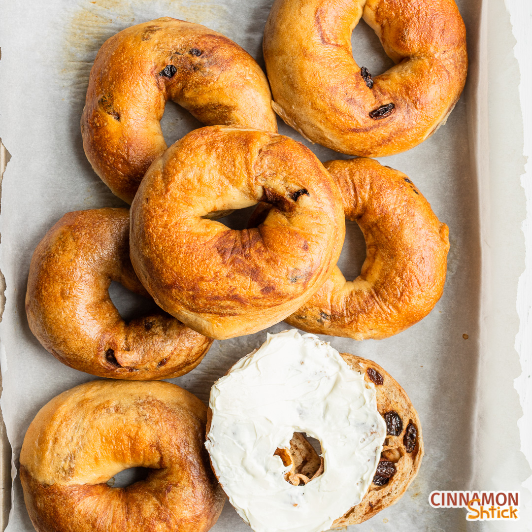 7 cinnamon raising bagels on parchment lined baking sheet, bunched together, with one sliced open and one-half with cream cheese spread on it