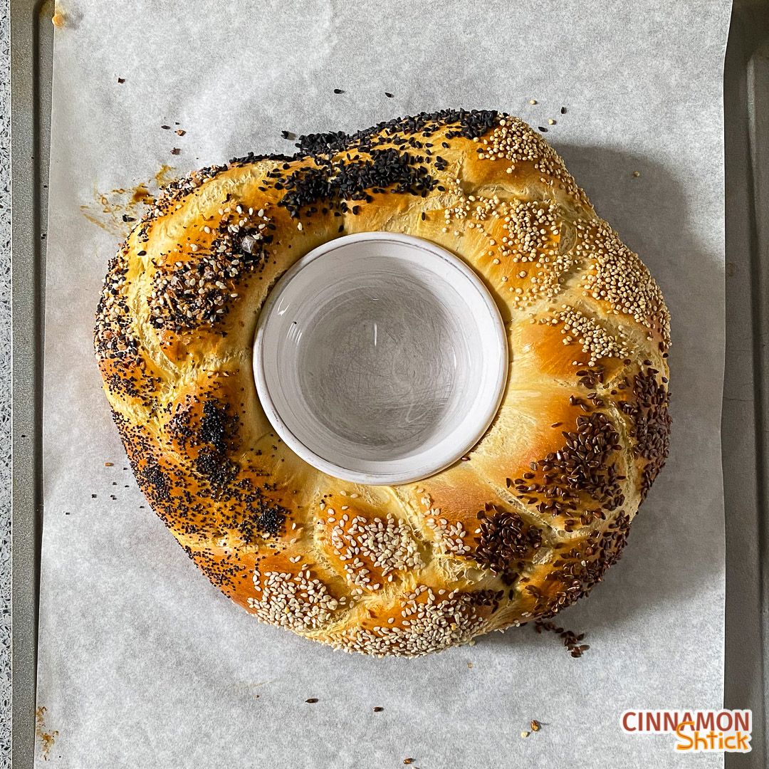 Three strand challah formed in a circle with a ramekin in the middle decorated with seeds just out of the oven.