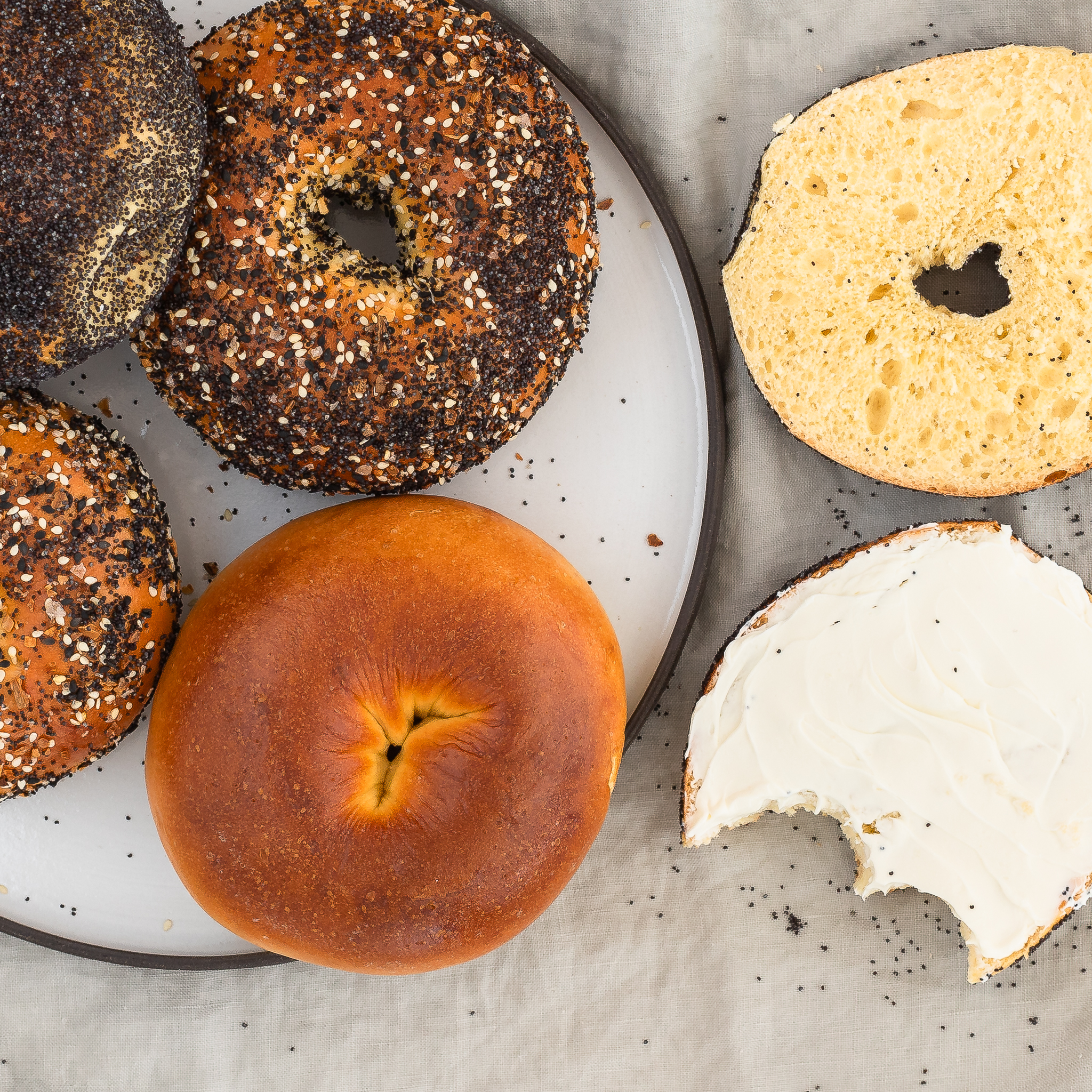 overhead view of an egg bagel cut in half, cream cheese on one side with a bite take out, and additional egg bagels on a plate to the left side