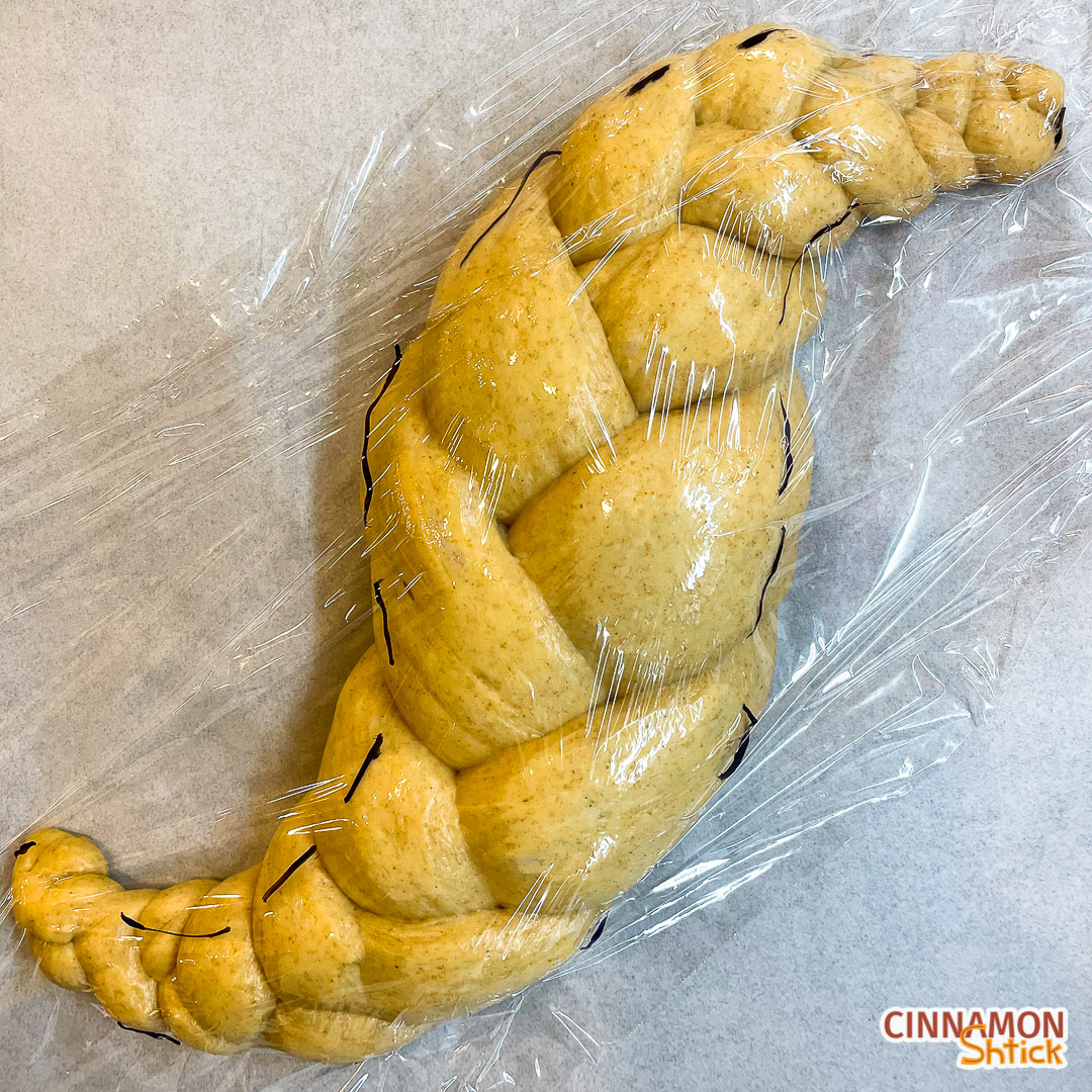 shaped sourdough challah with plastic wrap on it and markings to show how it has increased in size