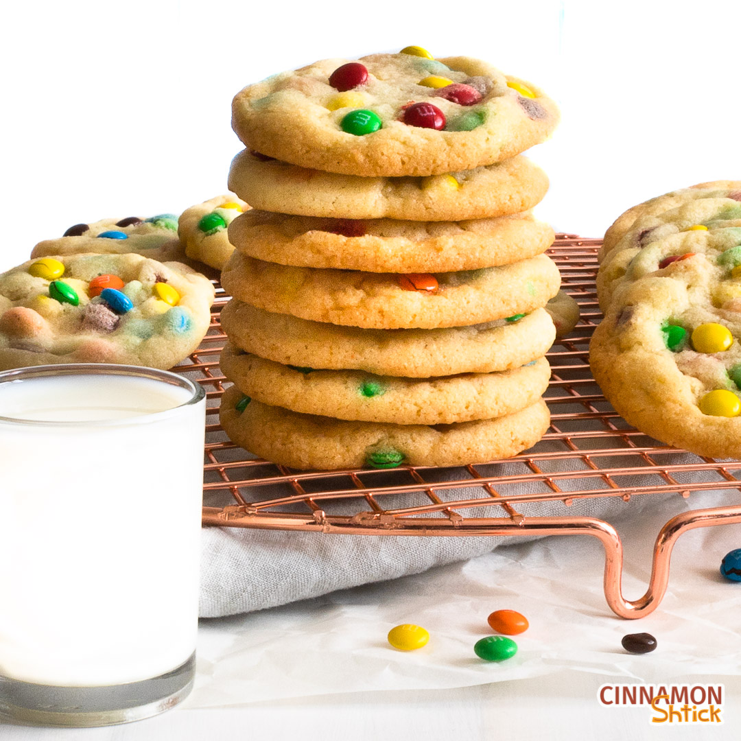 Stack of M&M cookies on cooling rack with glass of milk in forefront