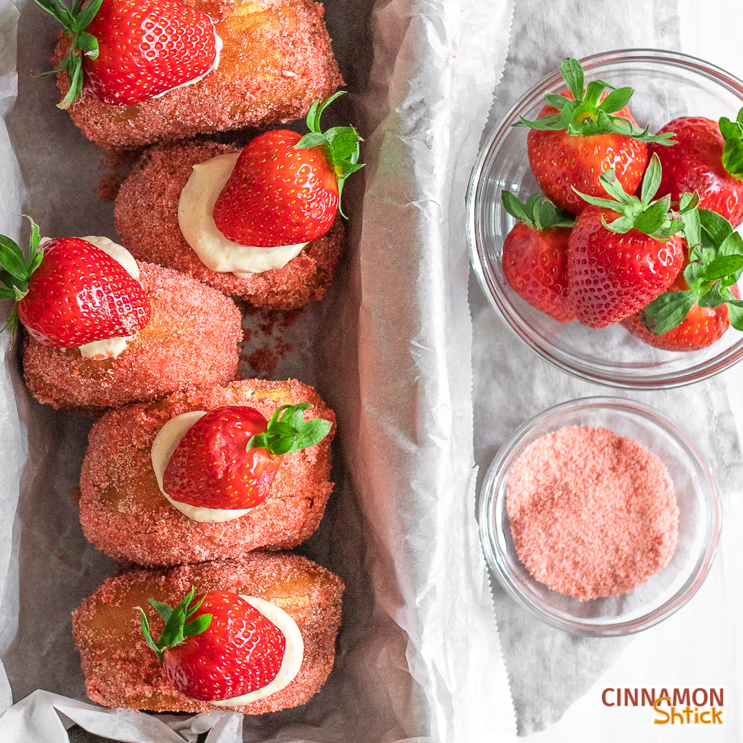 Strawberry donuts sitting upright in a loaf pan. To the right in the back are strawberries in a bowl in front is a bowl of strawberry sugar.