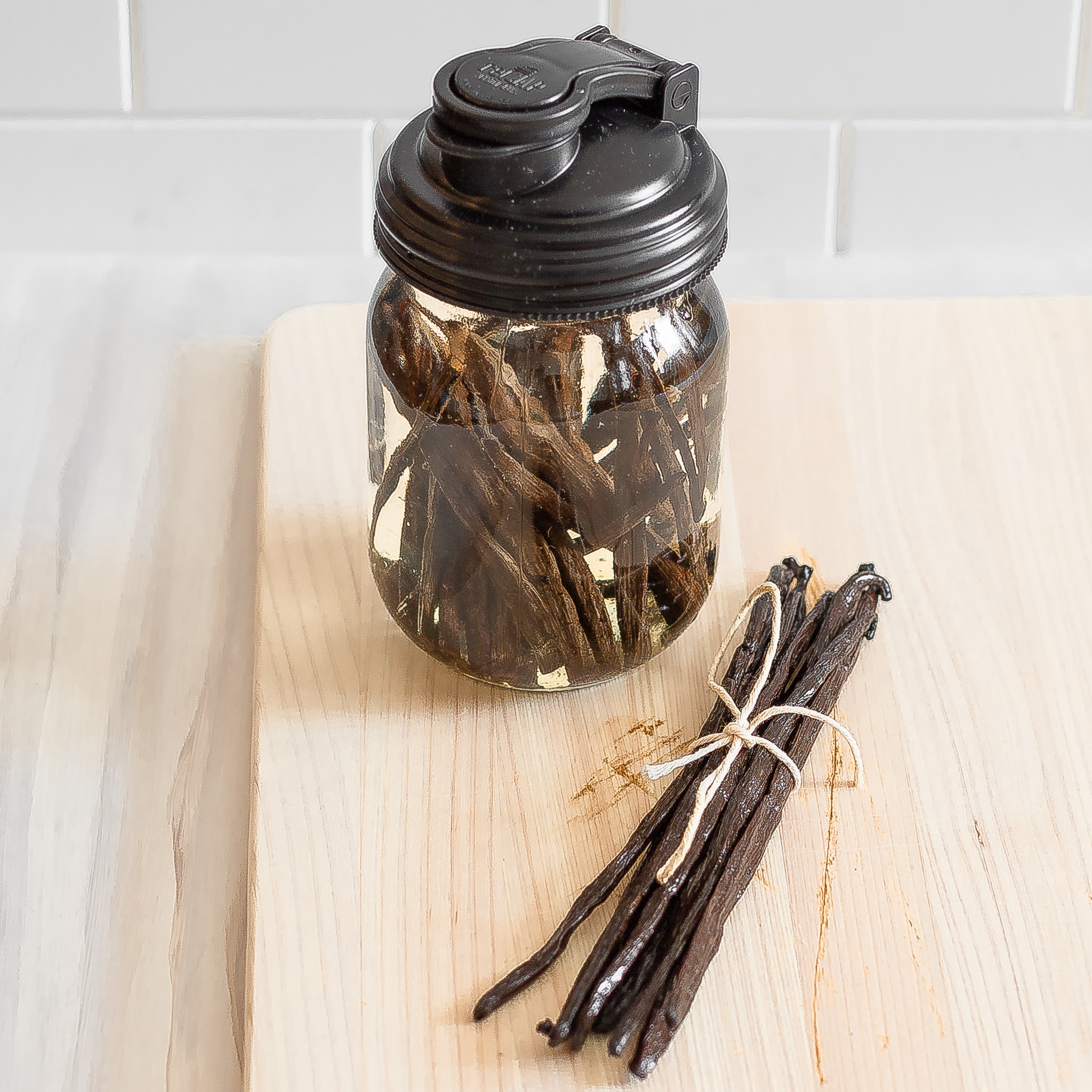 bunch of vanilla beans tied together in forefront with a mason jar closed with a re-cap filled with vanilla beans and vodka in upper left