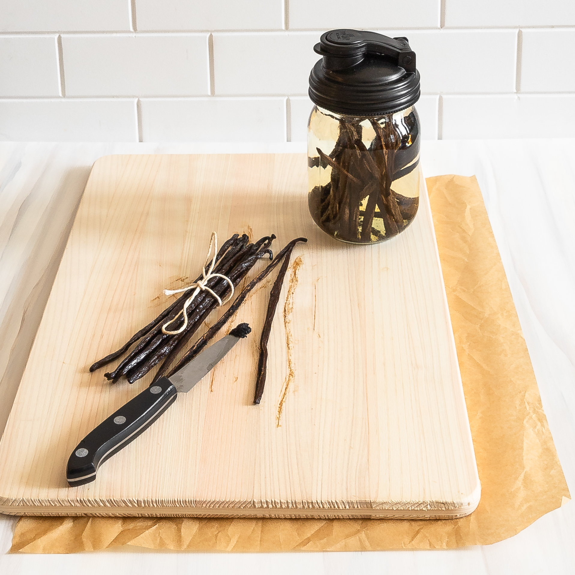 mason jar filled with alcohol and vanilla beans in the right background on a cutting board with vanilla beans and a paring knife in the foreground on the cutting board