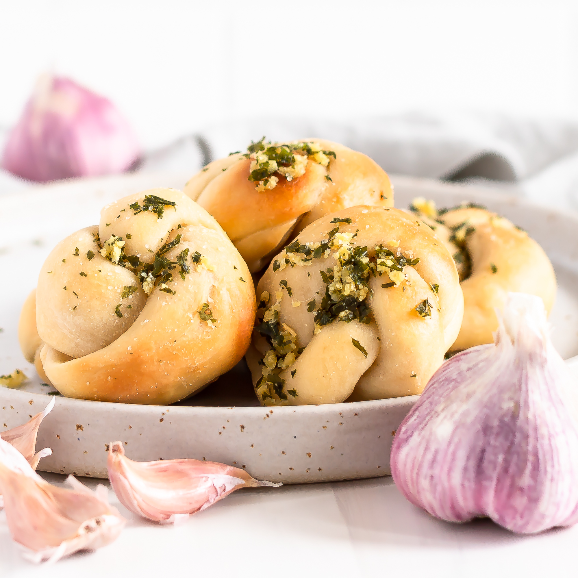 garlic knots on a plate with whole purple garlic and garlic cloves in the forefront and background