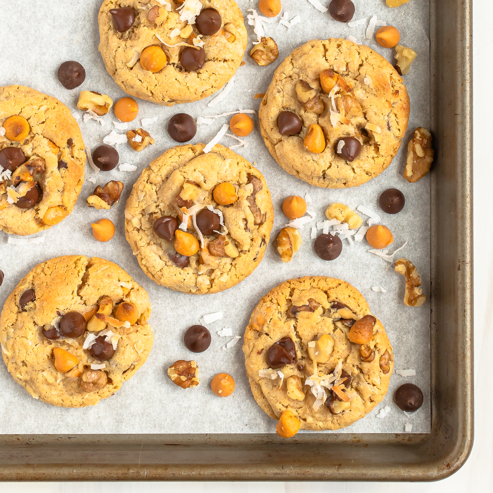 overhead view of magic bar cookies on parchment paper in a baking pan with walnuts, chocolate chips, butterscotch chips and coconut in between the cookies for decoration