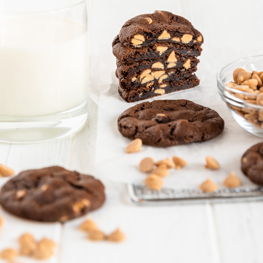 stack of chocolate peanut butter chip cookies sliced in half next to glass of milk with a couple cookies in front and scattered peanut butter chips