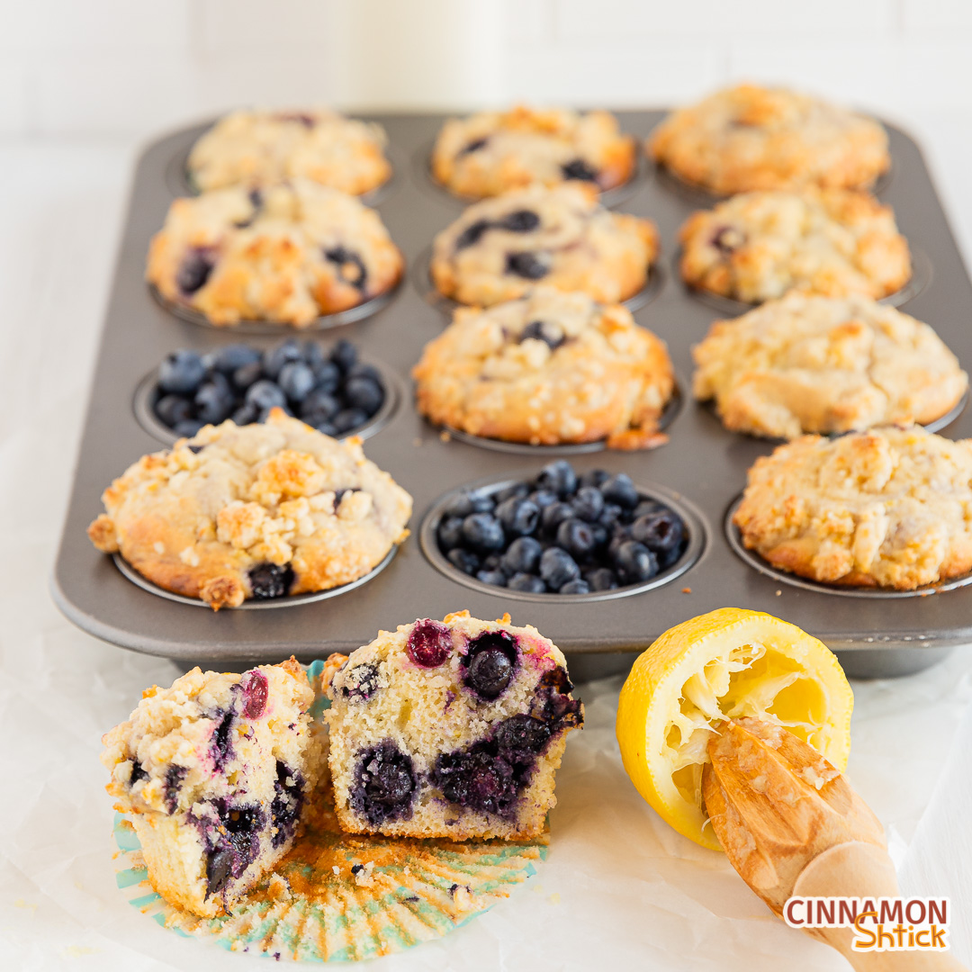 45 degree angle shot of a muffin cut in half next to a juiced lemon and a juice with a 12-tray of muffins behind them