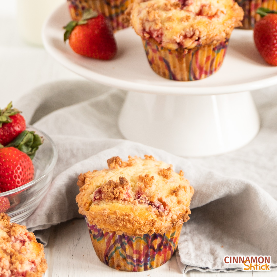 strawberry muffin in forefront with small cake standing holding more muffins and strawberries in background and bowl of strawberries off to side