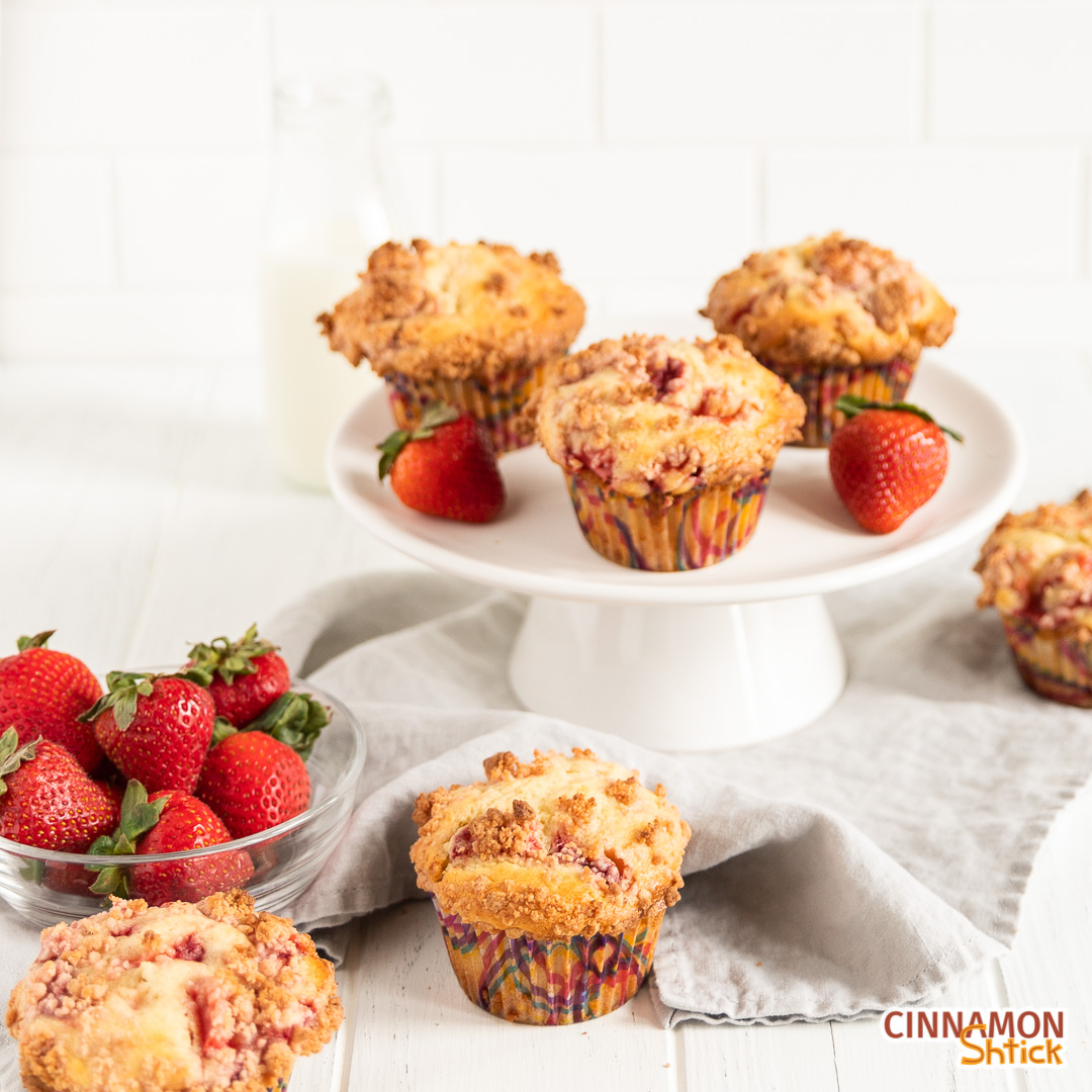 three strawberry muffins on a small cake stand with a couple more muffins in background, one in the forefront, and a bowl of strawberries in the left forefront