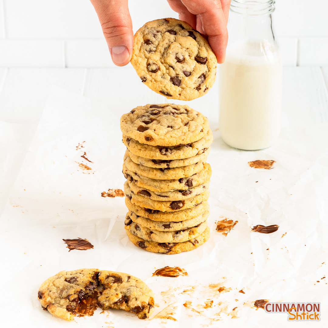 stack of vegan chocolate chip cookies with a hand taking the top one. In the foreground to the left is a cookie with a bite taken out and in the background to the right is a bottle of milk