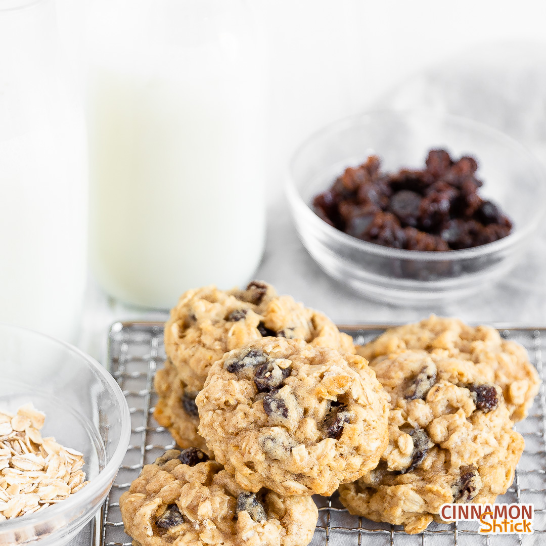 five vegan oatmeal vegan raisin cookies piled on a cooling rack with a small bowl of oatmeal to the left, two bottle of oat milk in the upper left, and a small bowl of raisins in the upper right