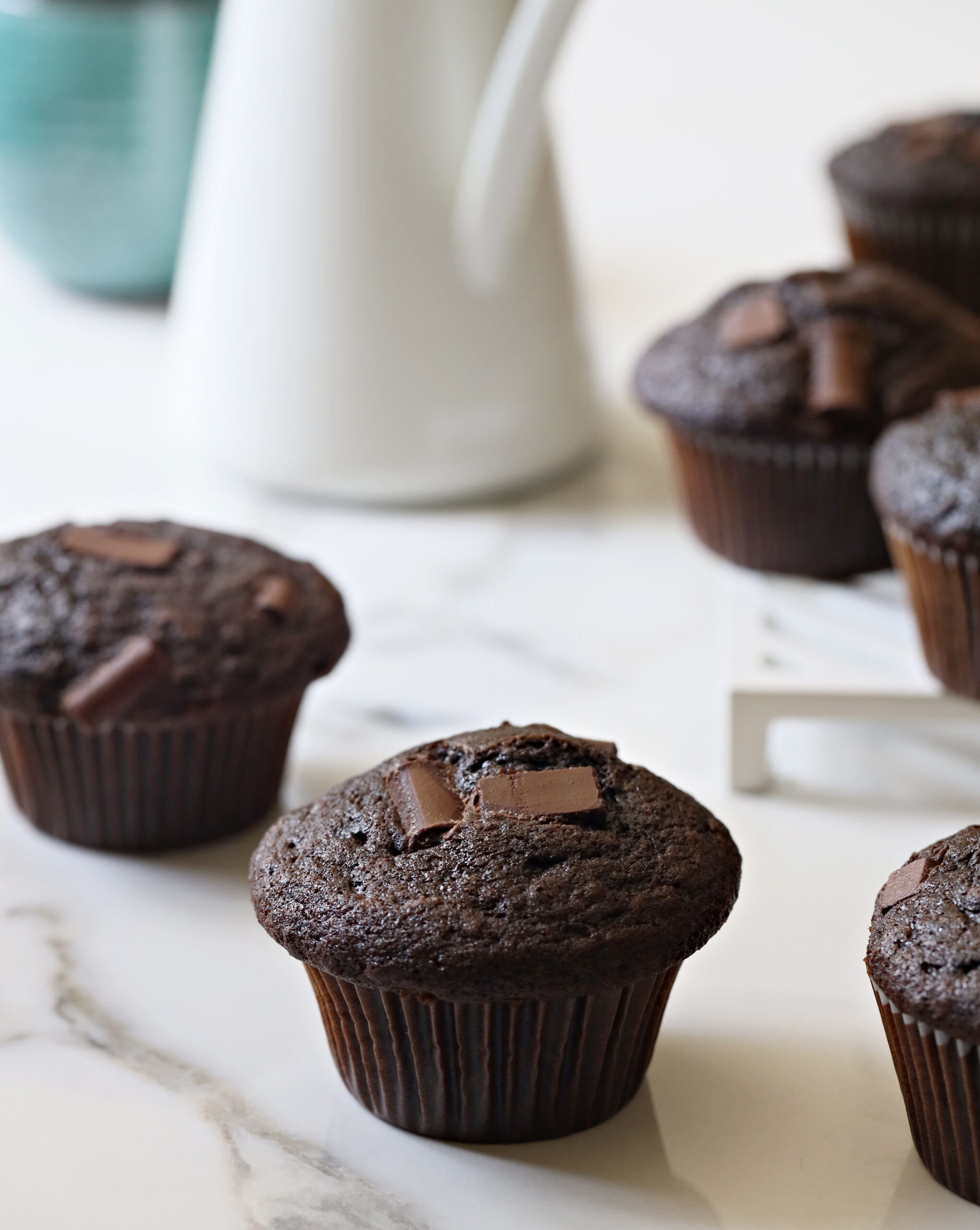 two bakery style chocolate muffins with white pitcher in background