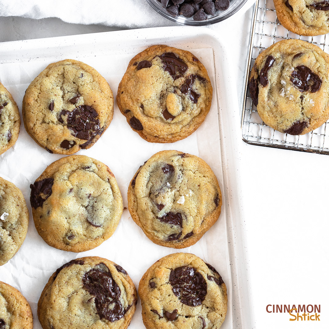 sourdough chocolate chip cookies on baking pan with a couple on a cooling rack and a bowl of chocolate chips