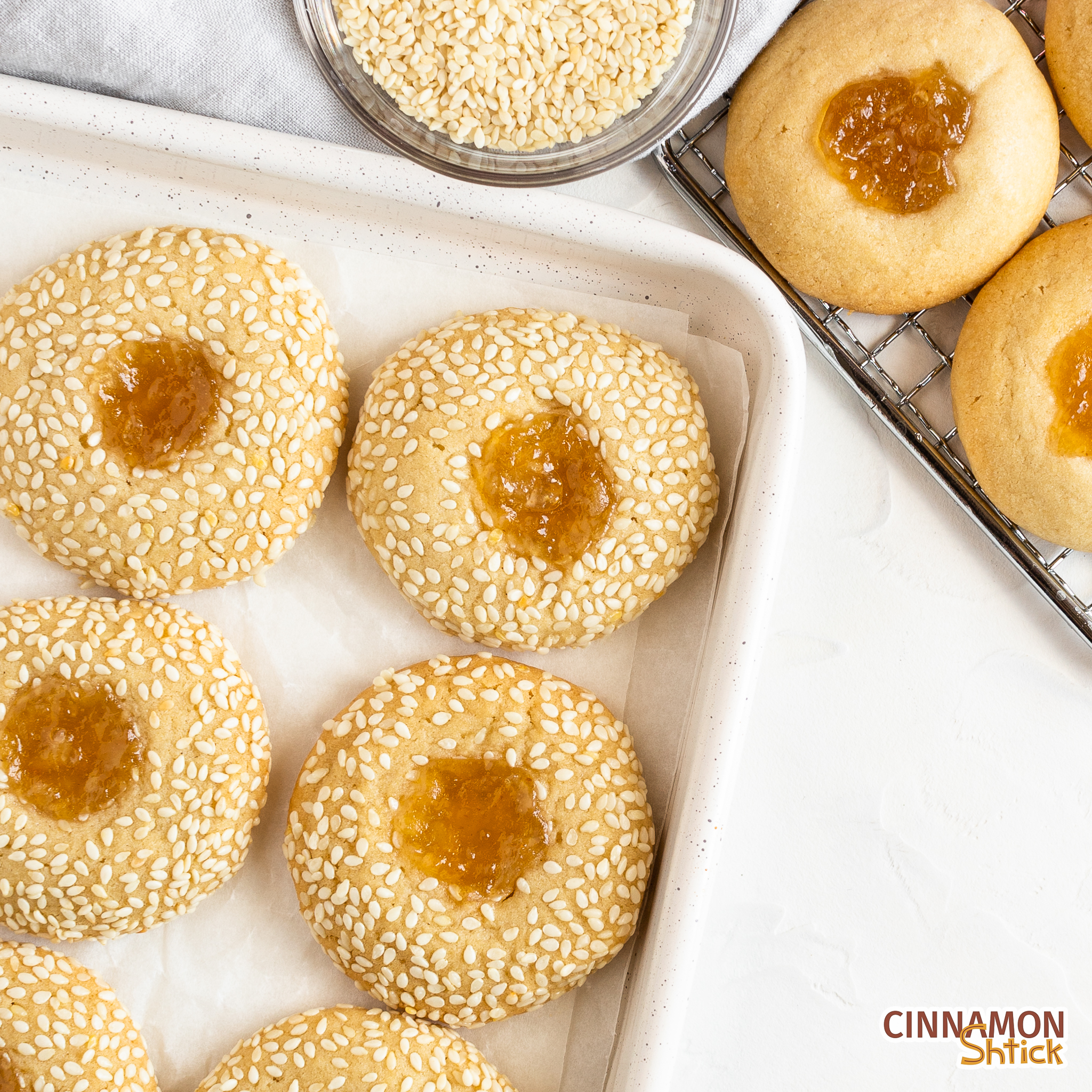 tahini thumbprint cookies in baking sheet with a couple on a cooling rack and a small bowl of sesame seeds