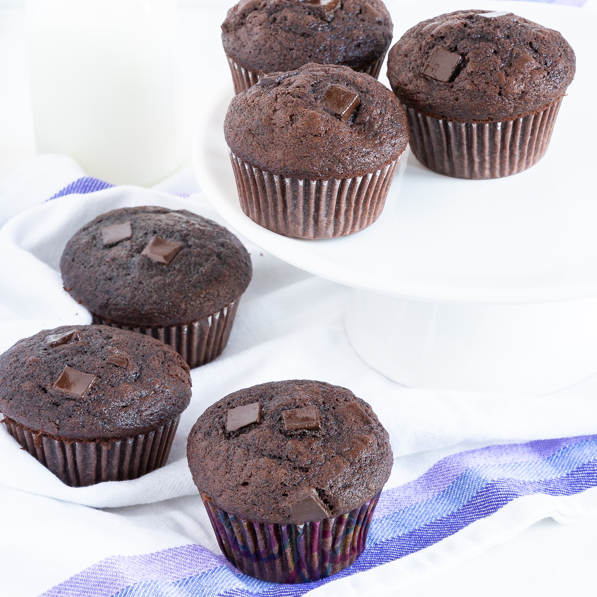 three bakery style chocolate muffins on a towel with three more on an elevated cake plate