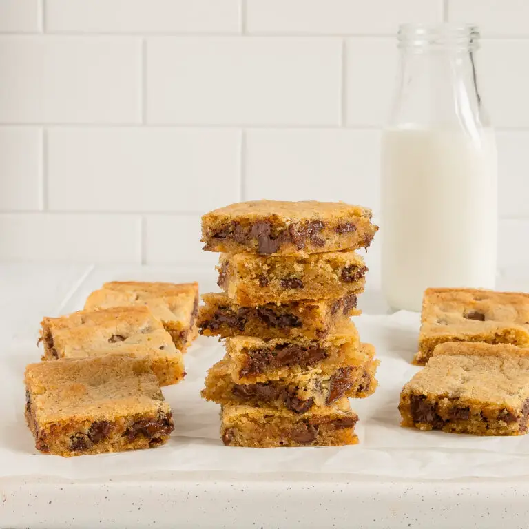 straight on shot of chocolate chip cookie bars stacked up on each other with some scattered in the background and a bottle of milk in the right background.