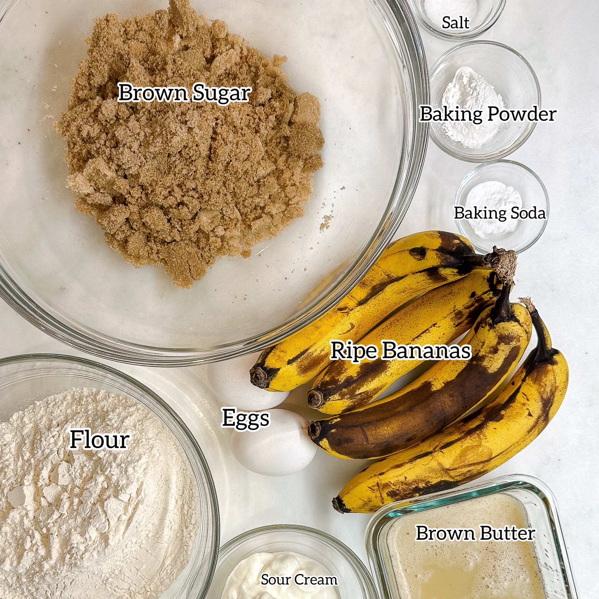 Overhead view of banana bread ingredients: brown sugar, flour, sour cream, brown butter, ripe bananas, baking soda, baking powder, and salt.