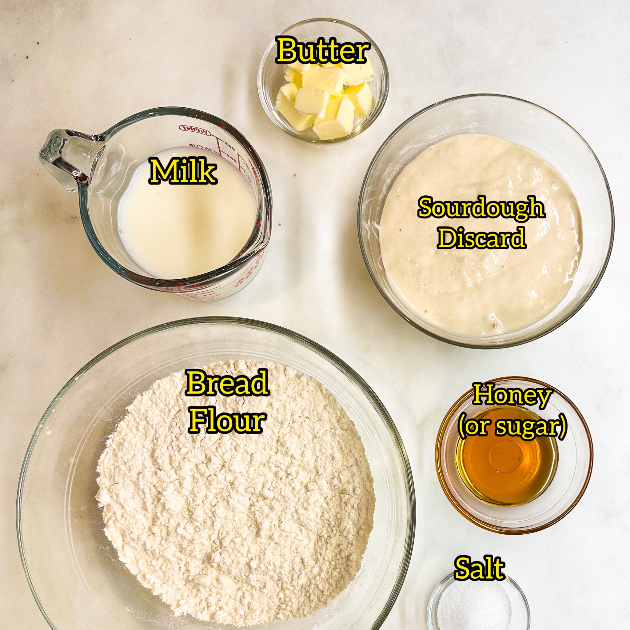 overhead shot of the ingredients in small bowls for sourdough discard pretzels. Clockwise from the top, butter, discard, honey, salt, bread flour and milk