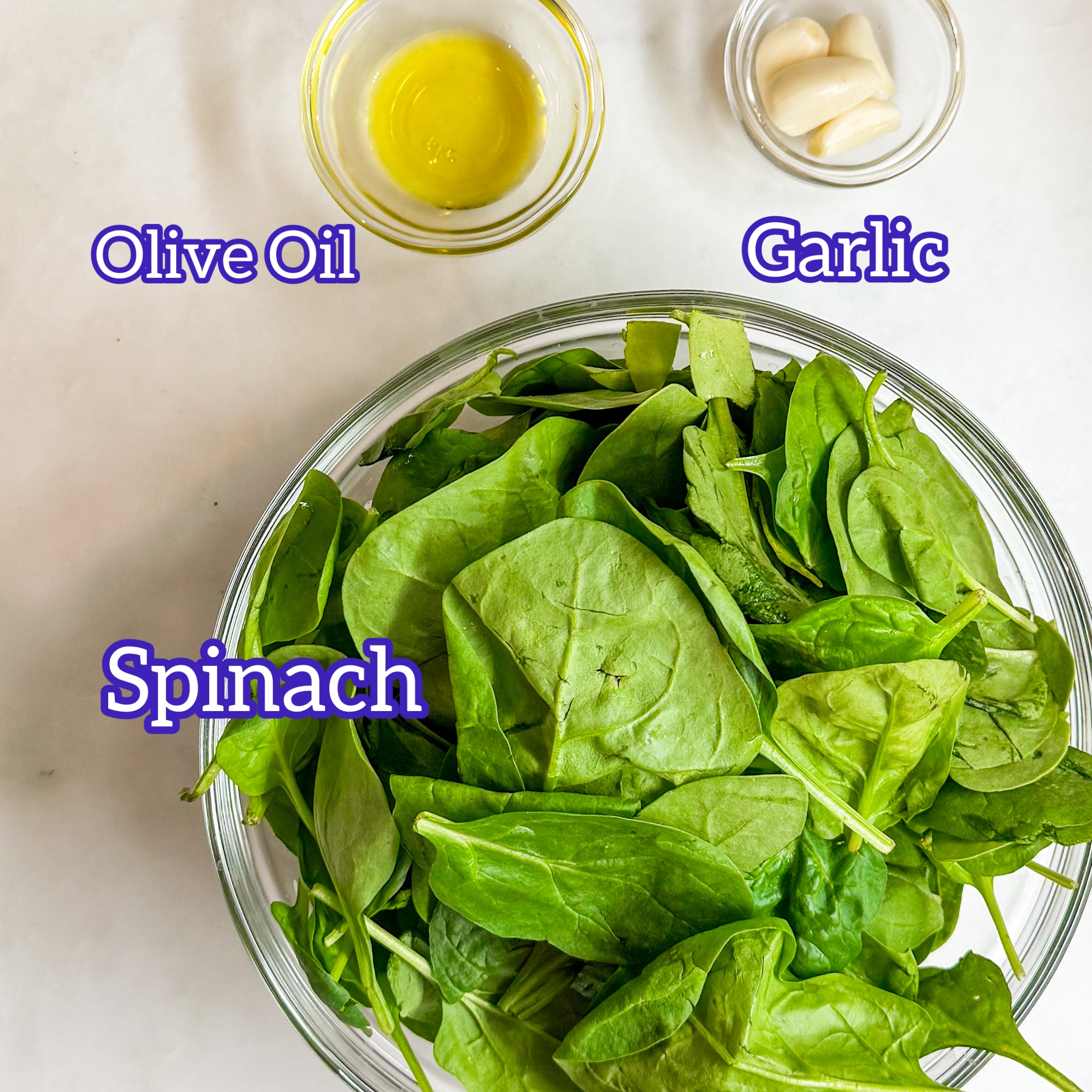 Overhead ov a bowl of spinach, a small bowl of olive oil and a small bowl with 4 cloves of garlic.