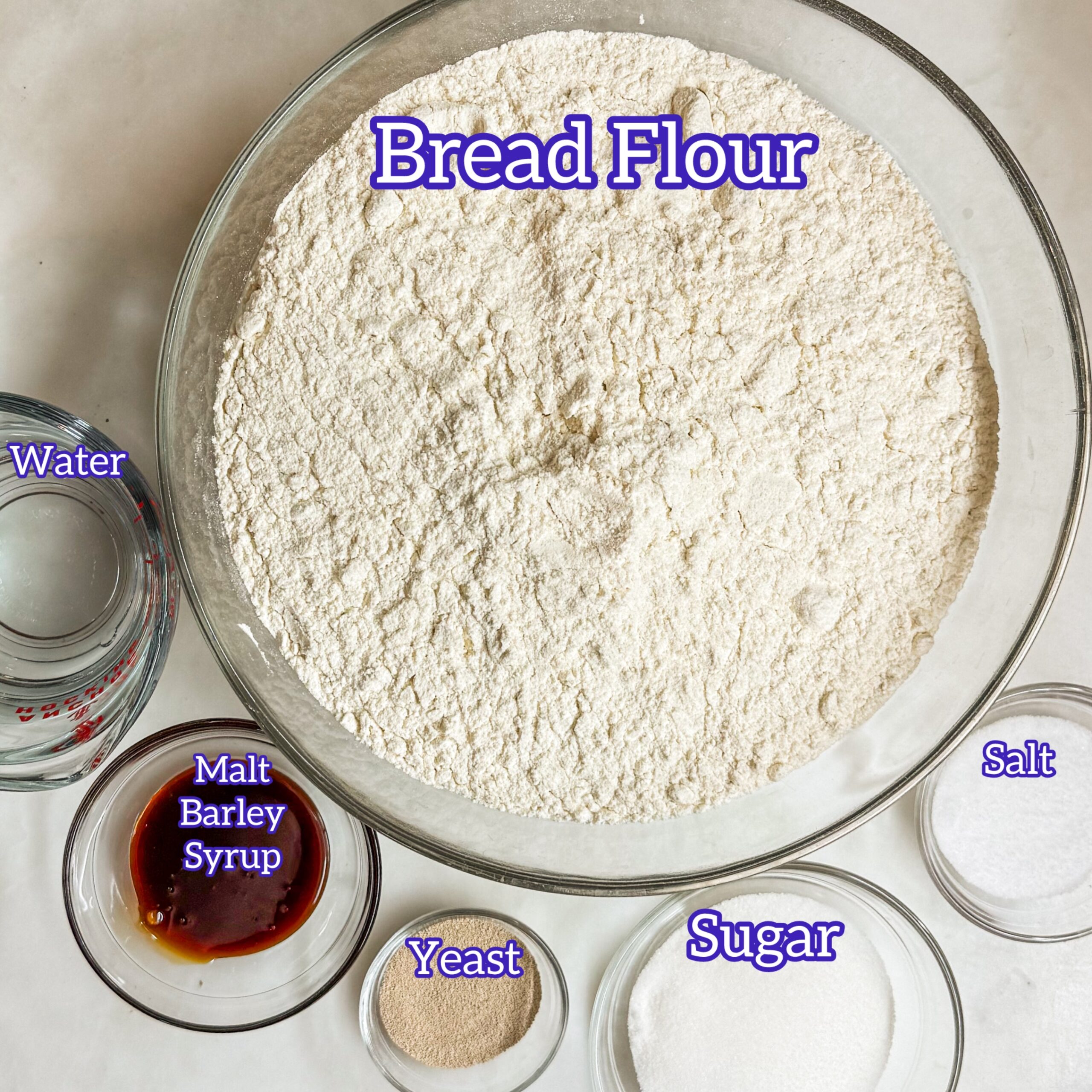 Overhead, in clockwise order, of a bowl of bread flour, a bowl of salt, a bowl of sugar, a bowl of yeast, a bowl of malt barley syrup and a bowl of water.