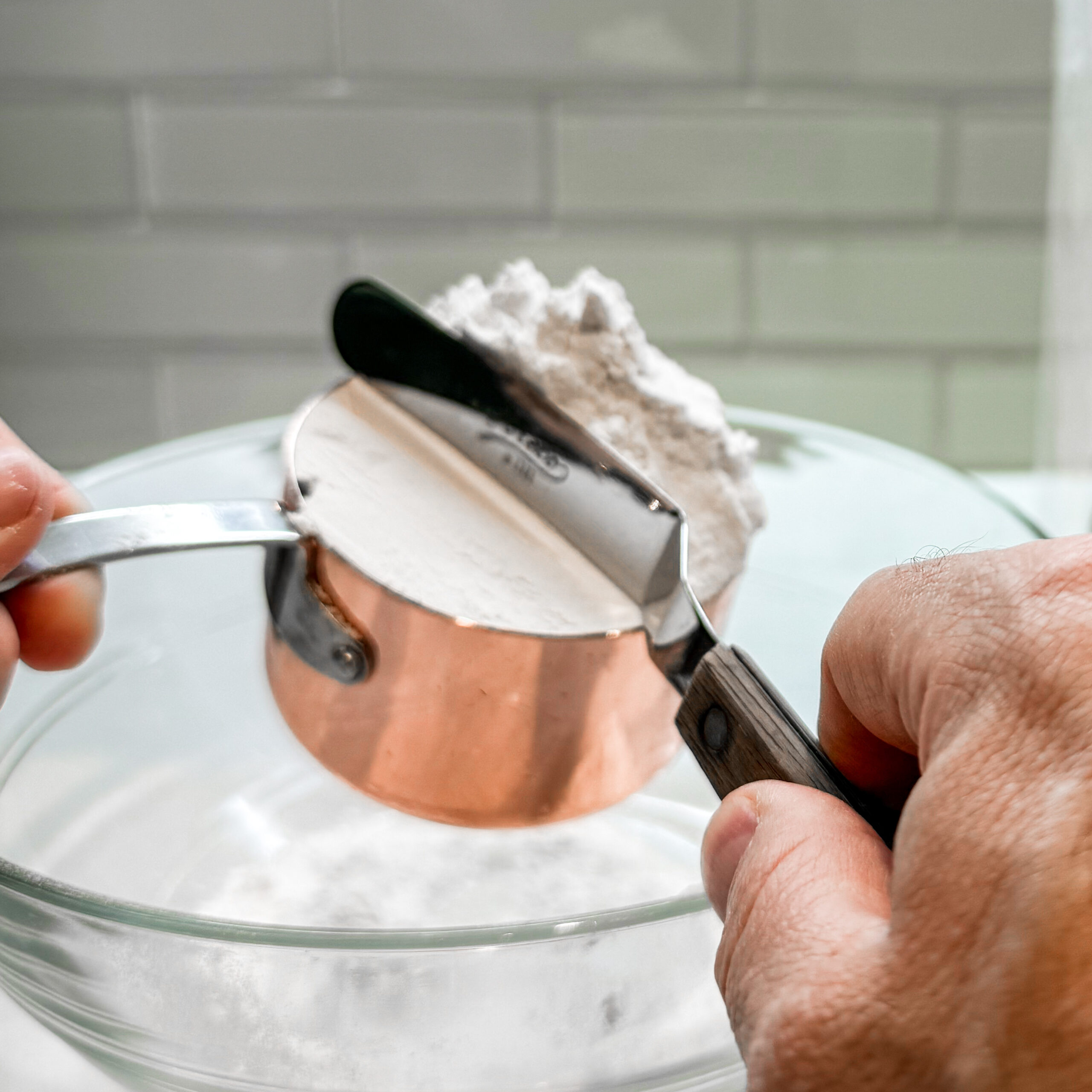 A metal spatula scraping the stop of the measuring cup leveling off the flour. 