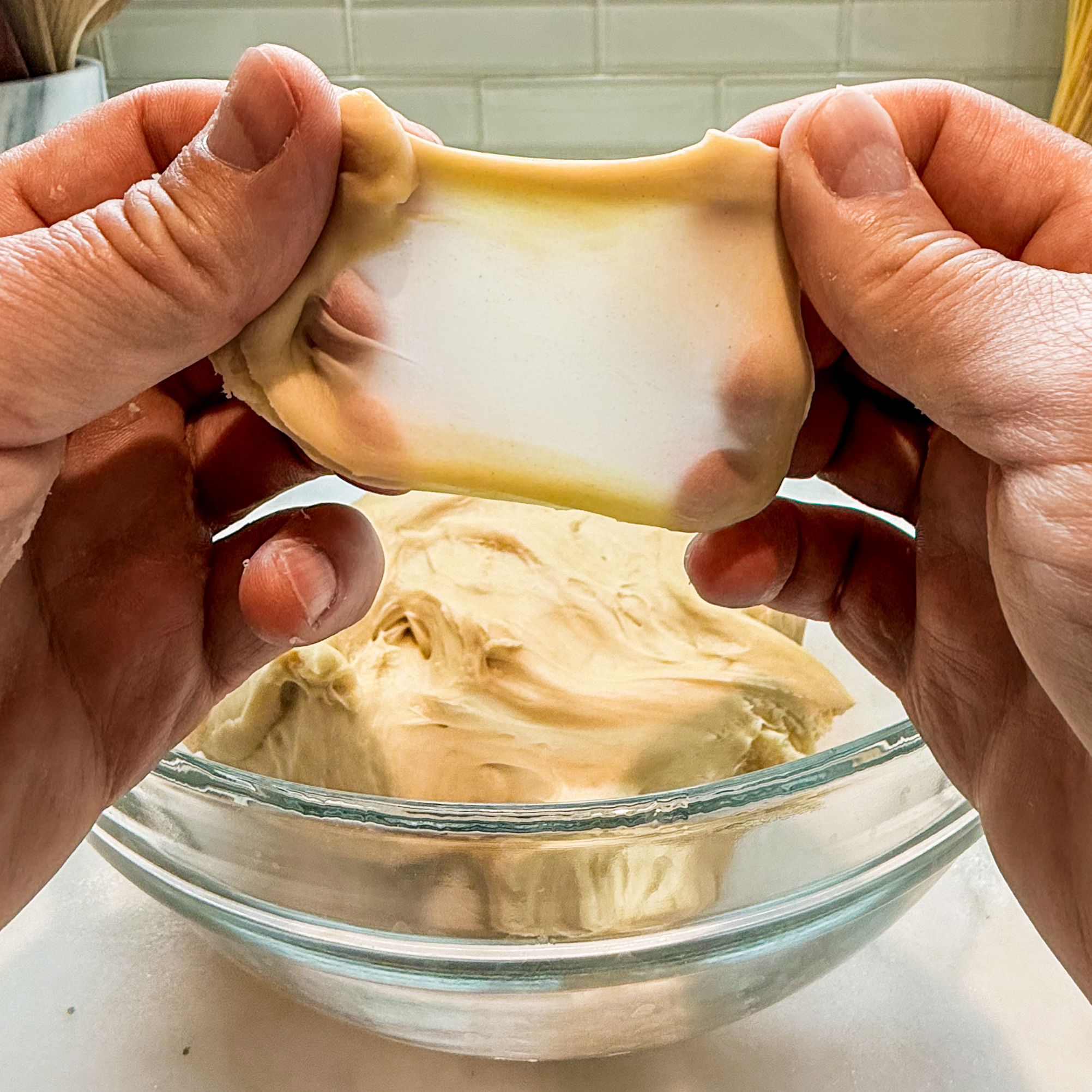 A piece of challah dough stretched between two hands showing the dough to be translucent.