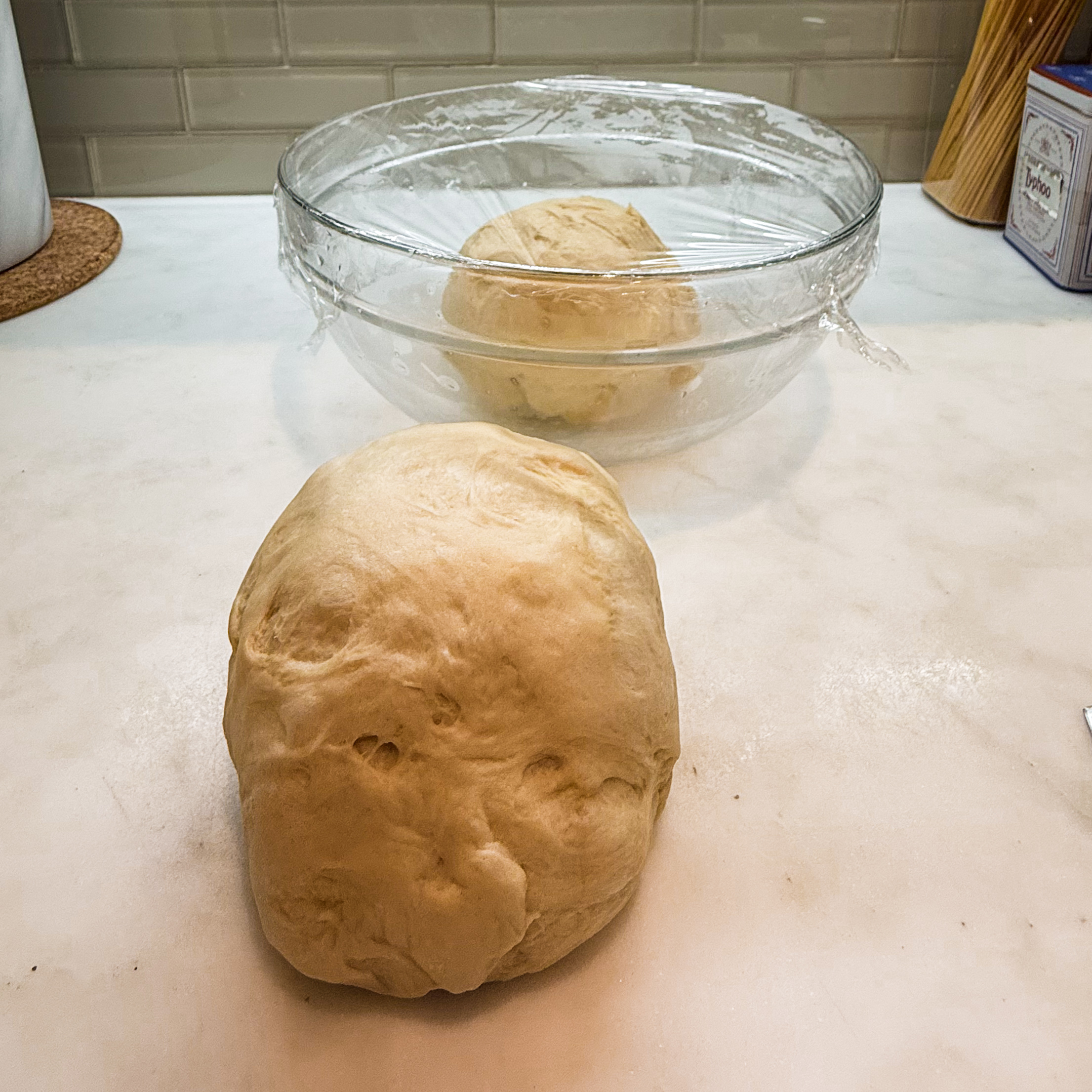 A ball of dough on a countertop with a bowl of dough covered with plastic wrap in the background.