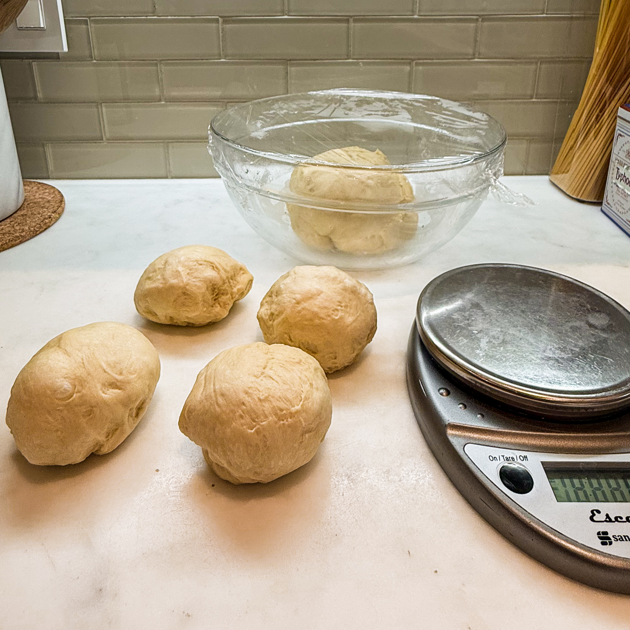 Four balls of challah dough with a portion of dough covered with plastic wrap in the background. 