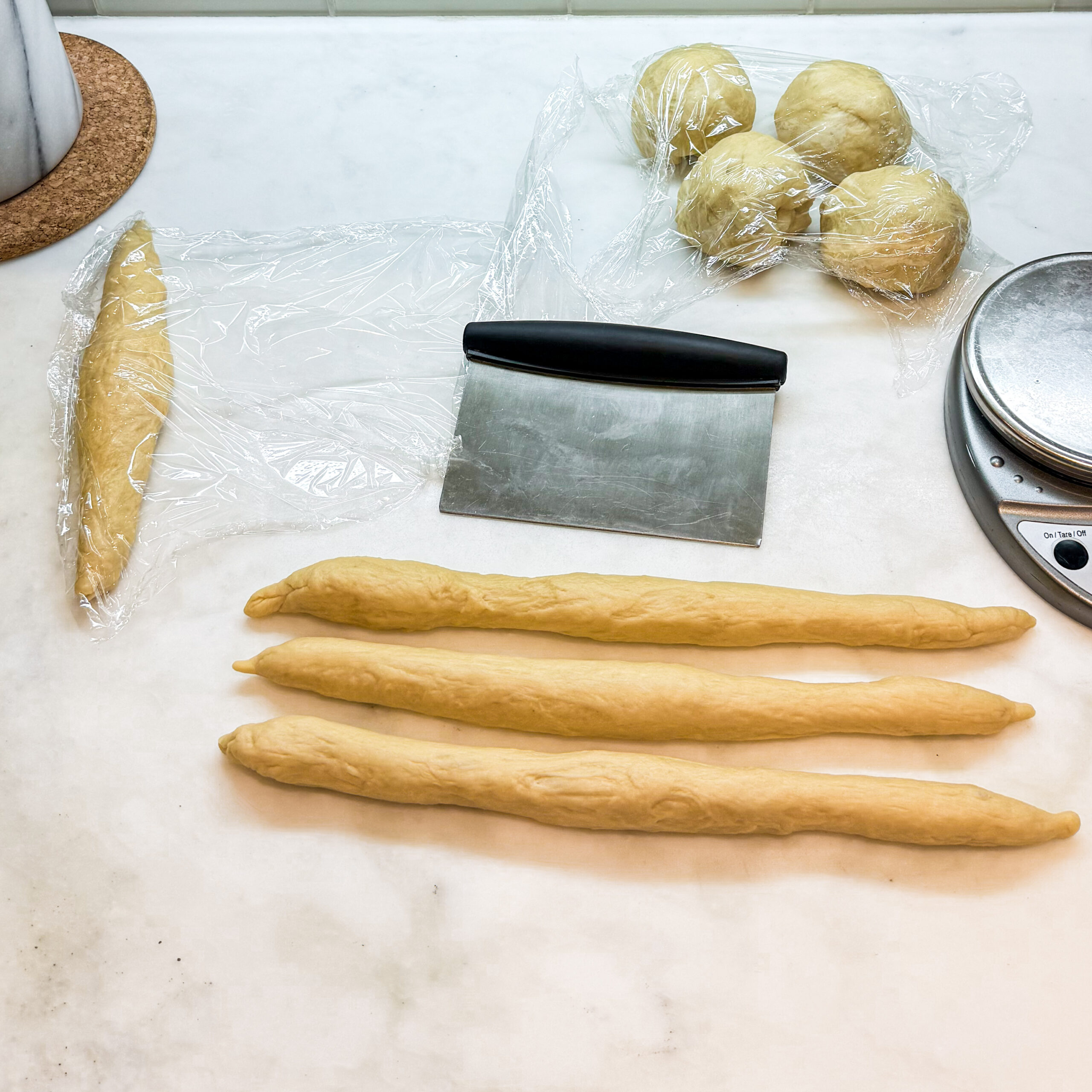 Three stands of dough rolled out with a fourth covered with plastic wrap and four dough balls in the background.