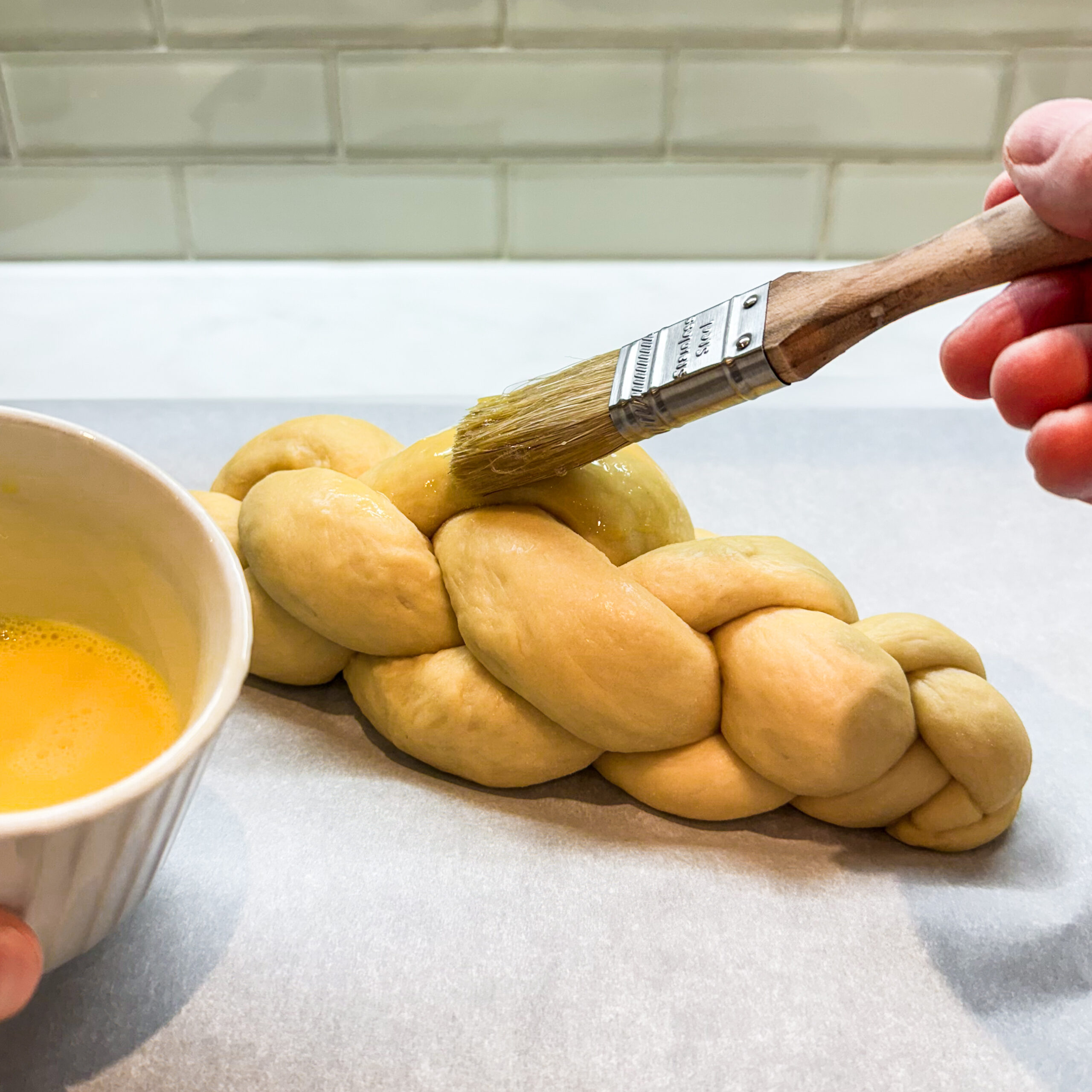 Pastry brush applying a beaten egg to a loaf of fully proofed challah.