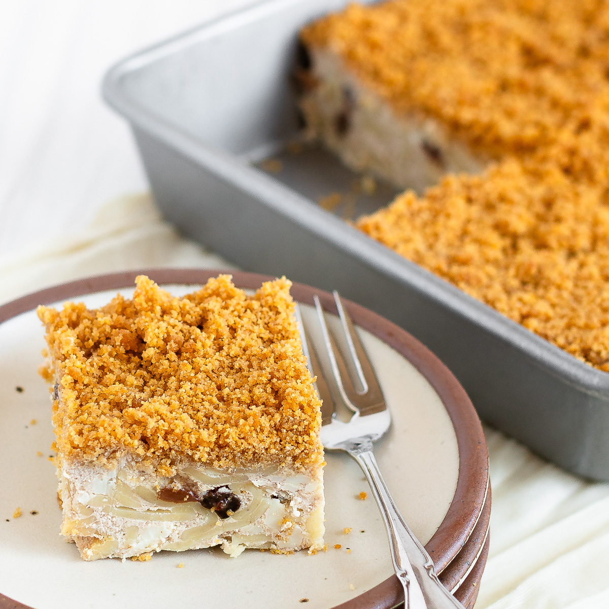 A piece of cheese kugel on a plate with a fork with the rest of the pan of kugel in the background.