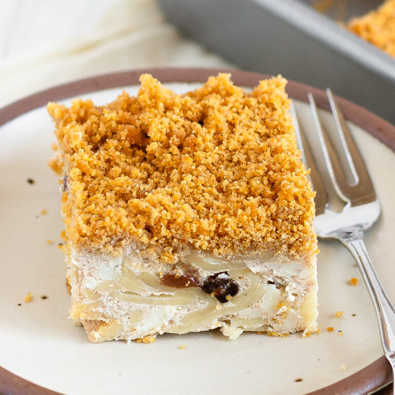 A piece of cheese kugel on a plate with a fork with the rest of the pan of kugel in the background.