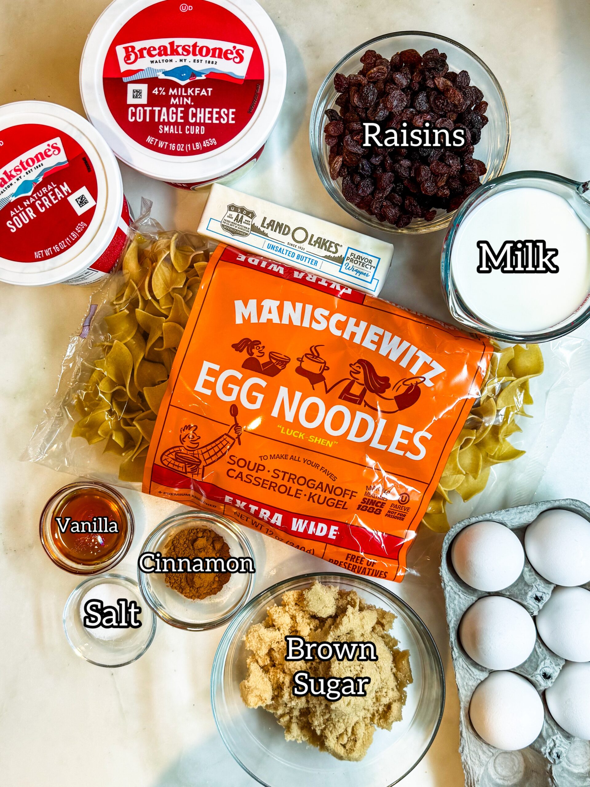 Overhead of kugel ingredients: sour cream, cottage cheese, raisins, butter, milk, noodles, eggs, brown sugar, cinnamon and vanilla extract.